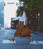 A woman in a white sweater and brown skirt crosses a city street at a crosswalk, carrying a large brown bag and wearing the Bed|Stü Karaoke lace-up chunky wooden platform shoes inspired by 70s shoe styles.
