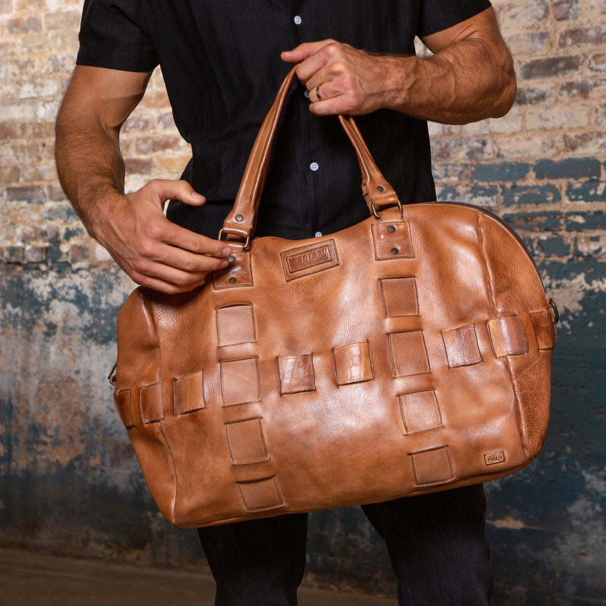 A man wearing a black shirt holds the Bed|Stü Keg, a large tan duffle featuring woven detailing, in front of a textured brick wall, showcasing the perfect travel bag crafted from premium vegetable tanned leather.