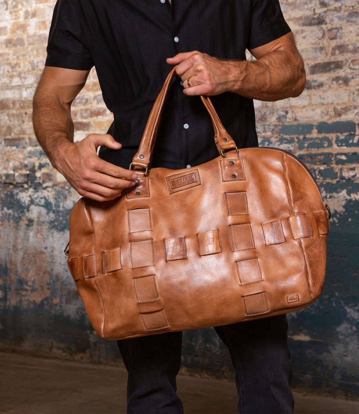 A man wearing a black shirt holds the Bed|Stü Keg, a large tan duffle featuring woven detailing, in front of a textured brick wall, showcasing the perfect travel bag crafted from premium vegetable tanned leather.