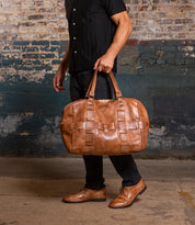 A man in a black shirt and pants holds the Bed|Stü Keg, a large tan leather duffle with woven details, standing against a textured brick and plaster wall.