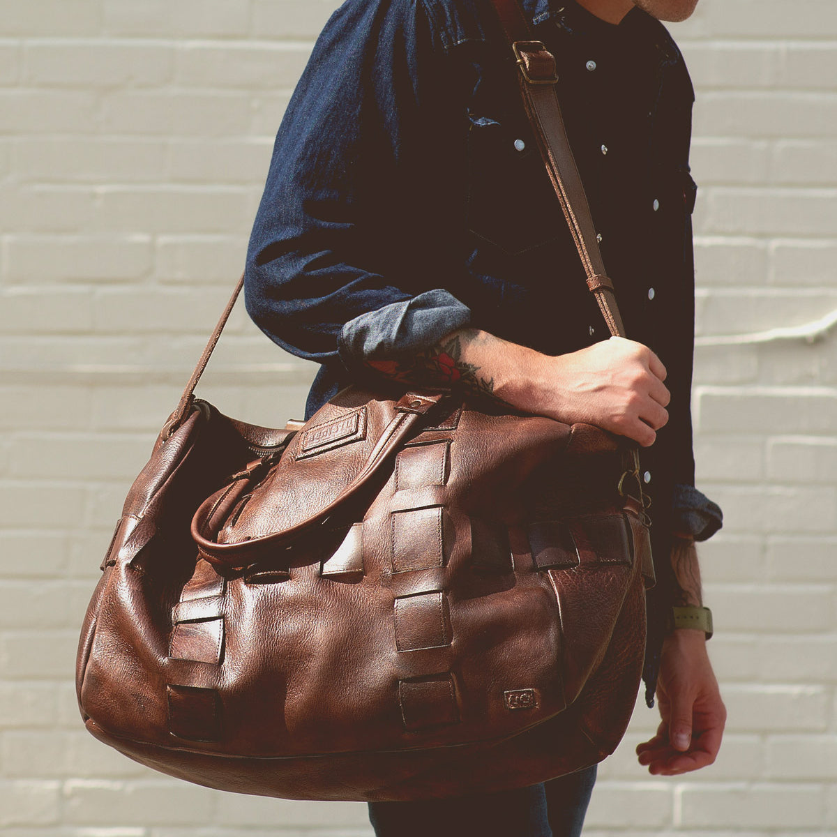 A person in a dark denim shirt carries the Bed|Stü Keg, a large vegetable tanned leather duffel bag, over their shoulder while standing in front of a light brick wall.