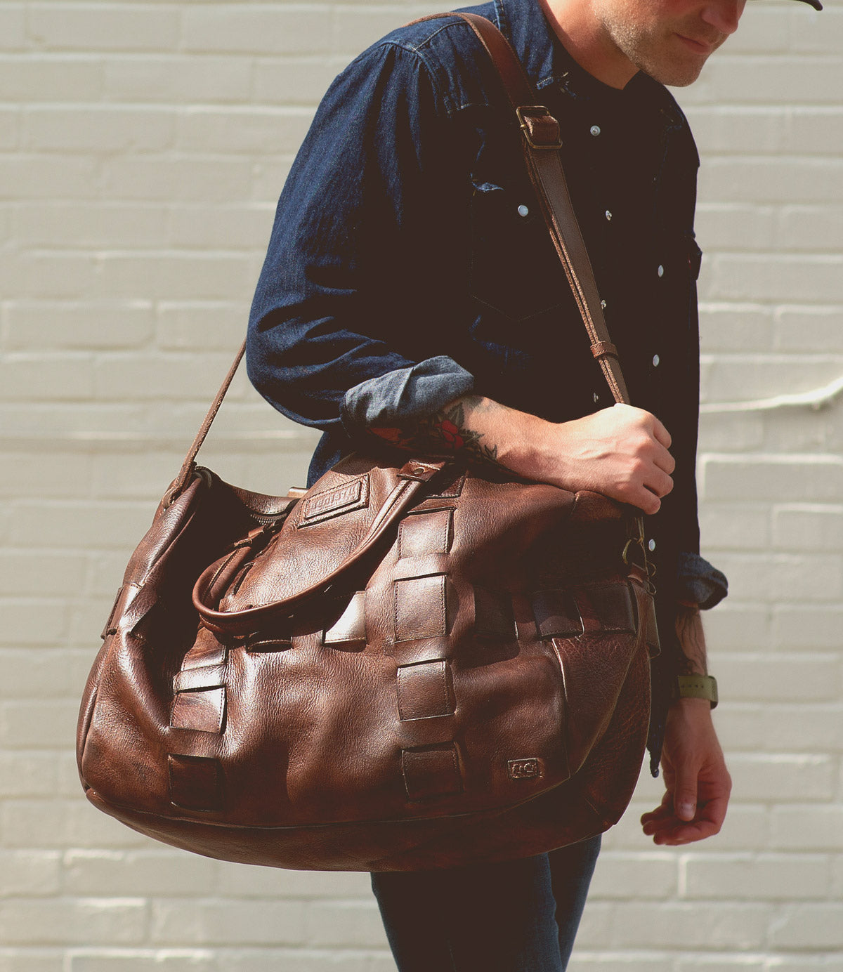 A person in a dark denim shirt carries the Bed|Stü Keg, a large vegetable tanned leather duffel bag, over their shoulder while standing in front of a light brick wall.