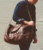 A person in a dark denim shirt carries the Bed|Stü Keg, a large vegetable tanned leather duffel bag, over their shoulder while standing in front of a light brick wall.