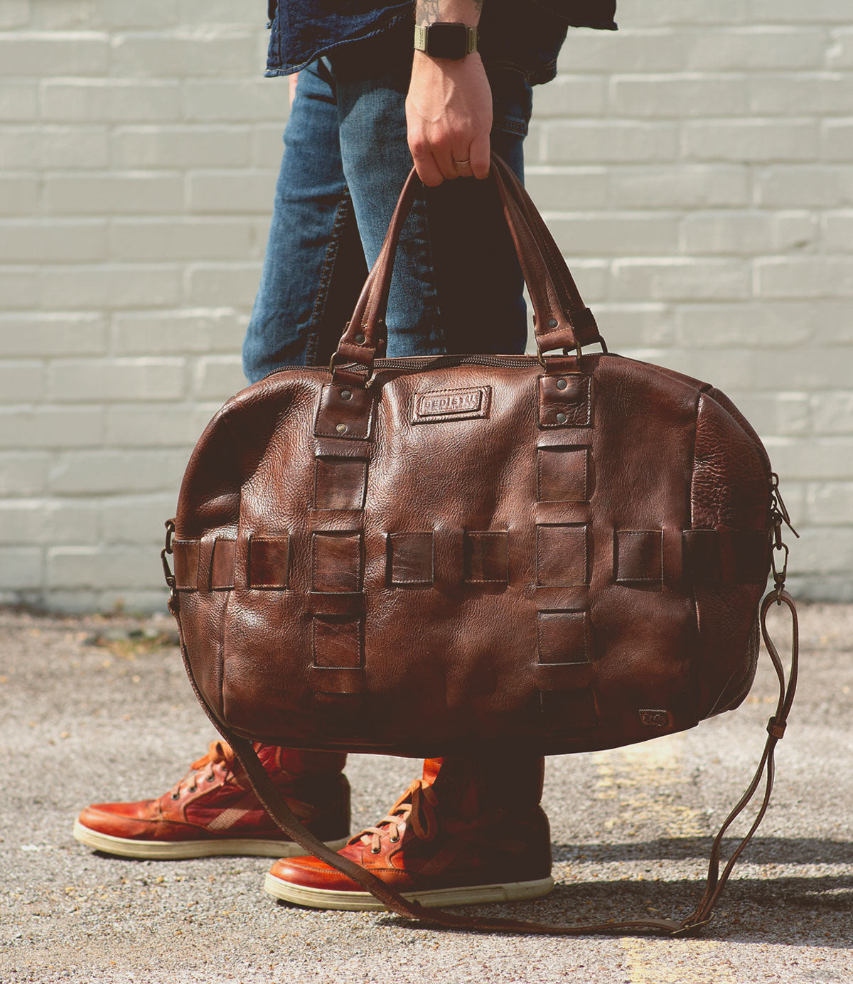 A person wearing jeans and red sneakers holds the Bed|Stü Keg, a large vegetable tanned leather duffle bag, by its handles while standing on a paved surface in front of a light-colored brick wall.