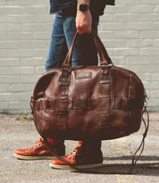 A person wearing jeans and red sneakers holds the Bed|Stü Keg, a large vegetable tanned leather duffle bag, by its handles while standing on a paved surface in front of a light-colored brick wall.