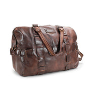 The Bed|Stü Keg is a brown leather duffel bag, thoughtfully crafted from vegetable tanned leather with a textured patchwork design. Featuring two top handles and a detachable shoulder strap, this stylish and durable travel bag is shown against a white background.