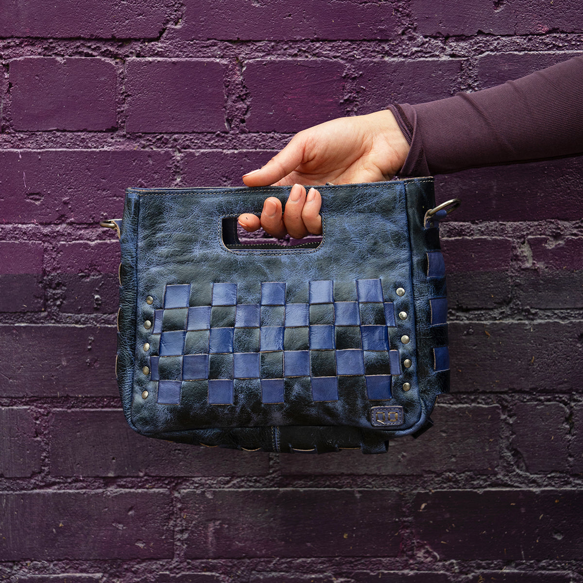 A hand holds the Bed|Stü Keiki clutch—a dark blue woven leather bag with a handle cutout and zip-top closure—in front of a textured purple brick wall.