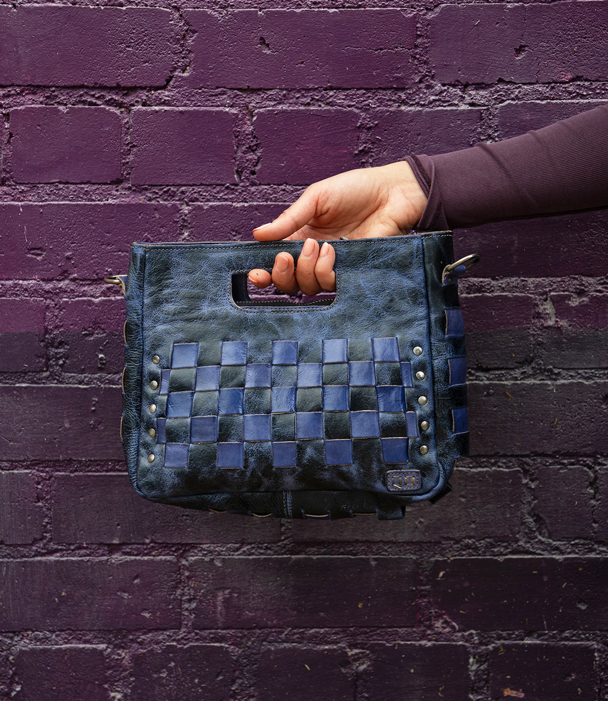 A hand holds the Bed|Stü Keiki clutch—a dark blue woven leather bag with a handle cutout and zip-top closure—in front of a textured purple brick wall.