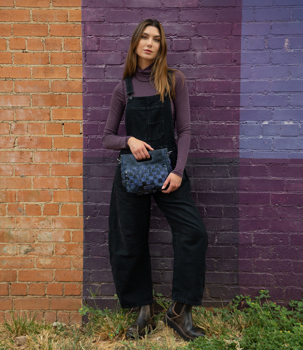 A woman stands against a brick wall painted in orange and purple, wearing a purple turtleneck, black overalls, boots, and holding the Bed|Stü Keiki blue checkerboard crossbody bag with an adjustable strap.