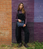 A woman stands against a brick wall painted in orange and purple, wearing a purple turtleneck, black overalls, boots, and holding the Bed|Stü Keiki blue checkerboard crossbody bag with an adjustable strap.