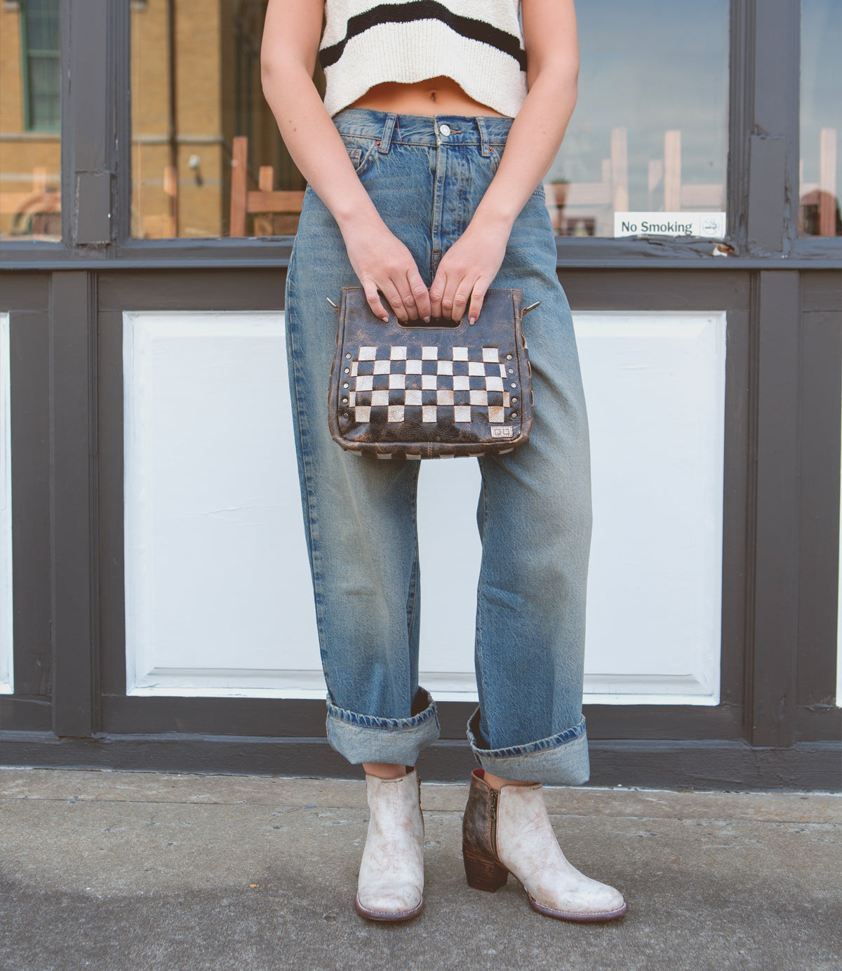 A person in cuffed jeans, a sleeveless crop top, and boots stands on a sidewalk holding the Bed|Stü Keiki—a sleek checkerboard crossbody bag with an adjustable strap and zip-top closure—in front of a doorway with a "No Smoking" sign.
