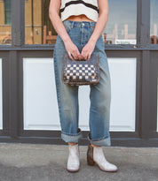 A person in cuffed jeans, a sleeveless crop top, and boots stands on a sidewalk holding the Bed|Stü Keiki—a sleek checkerboard crossbody bag with an adjustable strap and zip-top closure—in front of a doorway with a "No Smoking" sign.