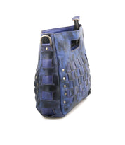 The Keiki by Bed|Stü is a blue leather checkerboard crossbody bag featuring a woven pattern, metal rivets, a zip-top closure, and a cut-out handle. Shown from a side angle against a white background, the bag also offers an adjustable strap for versatile wear.
