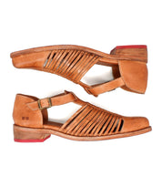 The Bed|Stü Kenna is a pair of tan leather sandals featuring a closed-toe loafer silhouette, woven side cutouts, adjustable ankle straps, low wooden heels, and distinctive red rubber heel accents.