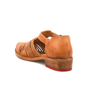The Bed|Stü Kenna is a brown leather closed-toe sandal featuring woven cutouts, a low heel, an adjustable ankle strap, and a red outsole accent, displayed from the rear on a white background.