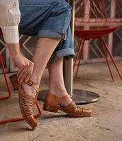 A person sitting on a red chair, wearing cuffed jeans, buckles the Bed|Stü Kenna—a brown closed-toe loafer featuring woven cutouts and an adjustable ankle strap—on their foot while crossing their other leg.