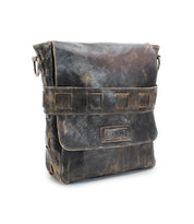 The Bed|Stü Kure is a rugged, distressed brown leather messenger bag with a flap closure and metal ring attachments, featuring a handwoven strap crafted from vegetable-tanned leather, shown against a white background.