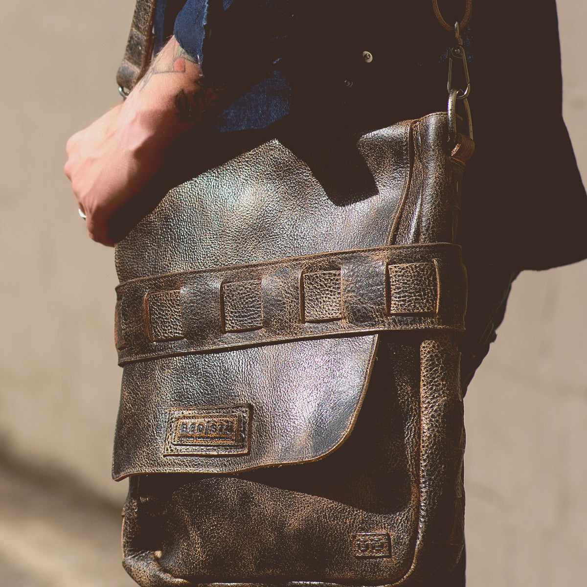 A person wearing a dark shirt holds the Bed|Stü Kure messenger bag, crafted from vegetable tanned distressed brown leather with a flap and visible stitching, standing against a neutral background.