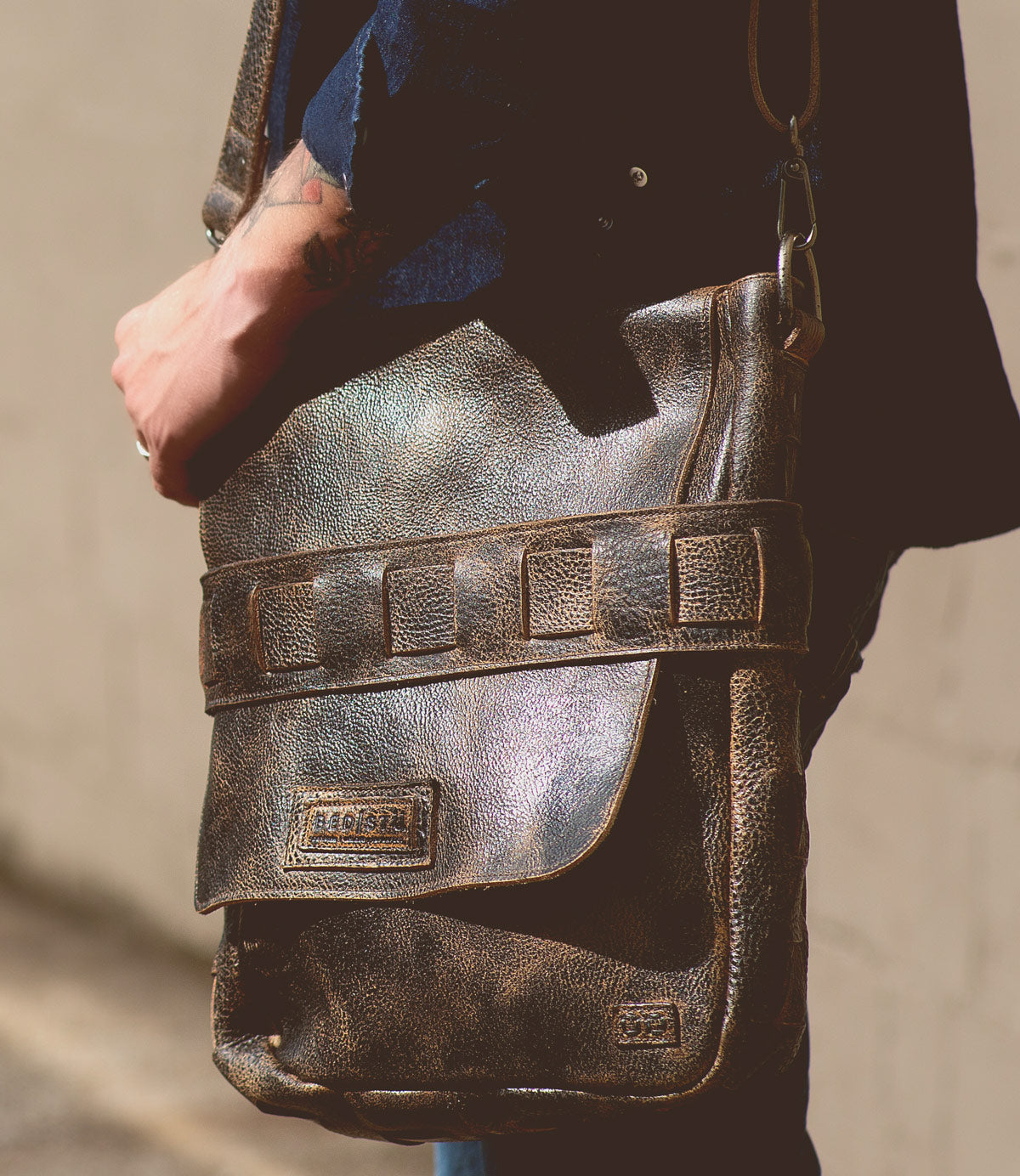 A person wearing a dark shirt holds the Bed|Stü Kure messenger bag, crafted from vegetable tanned distressed brown leather with a flap and visible stitching, standing against a neutral background.