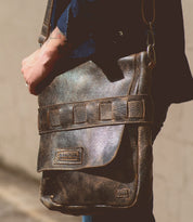 A person wearing a dark shirt holds the Bed|Stü Kure messenger bag, crafted from vegetable tanned distressed brown leather with a flap and visible stitching, standing against a neutral background.