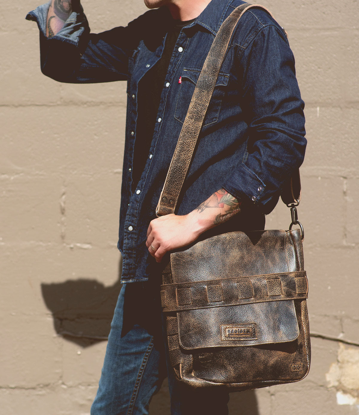 A person in a denim shirt and jeans carries the Bed|Stü Kure leather messenger bag with a handwoven strap, standing against a beige wall.