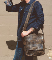 A person in a denim shirt and jeans carries the Bed|Stü Kure leather messenger bag with a handwoven strap, standing against a beige wall.