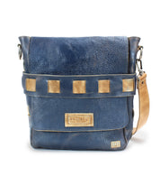 The Kure by Bed|Stü is a blue leather crossbody bag featuring a tan adjustable handwoven strap, square metal accents, and the Bed|Stü label on the front. Crafted from vegetable tanned leather, this distinctive accessory combines style and durability for everyday use.