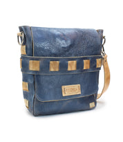 The Kure messenger bag by Bed|Stü is crafted from blue leather with tan square accents, featuring a vegetable-tanned leather shoulder strap and a front flap adorned with a stitched Bed|Stü label. The bag is displayed on a white background.