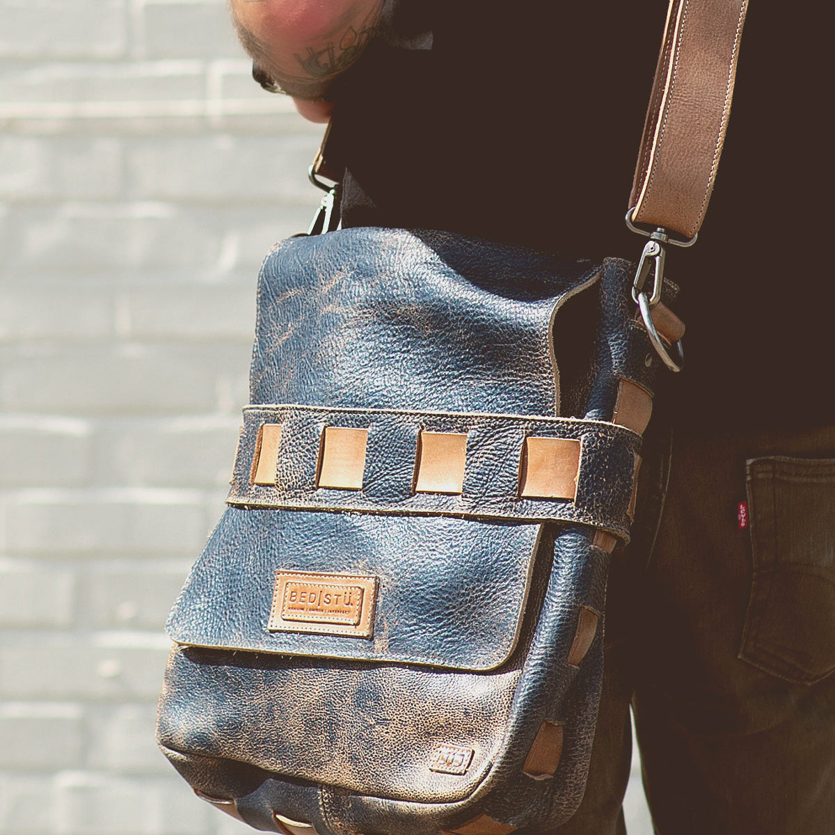 A person wearing the Bed|Stü Kure, a brown vegetable tanned leather messenger bag with tan accents and a handwoven strap, stands against a light-colored brick wall.
