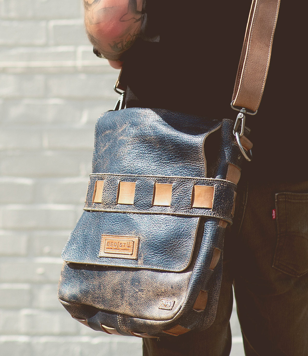 A person wearing the Bed|Stü Kure, a brown vegetable tanned leather messenger bag with tan accents and a handwoven strap, stands against a light-colored brick wall.