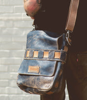 A person wearing the Bed|Stü Kure, a brown vegetable tanned leather messenger bag with tan accents and a handwoven strap, stands against a light-colored brick wall.