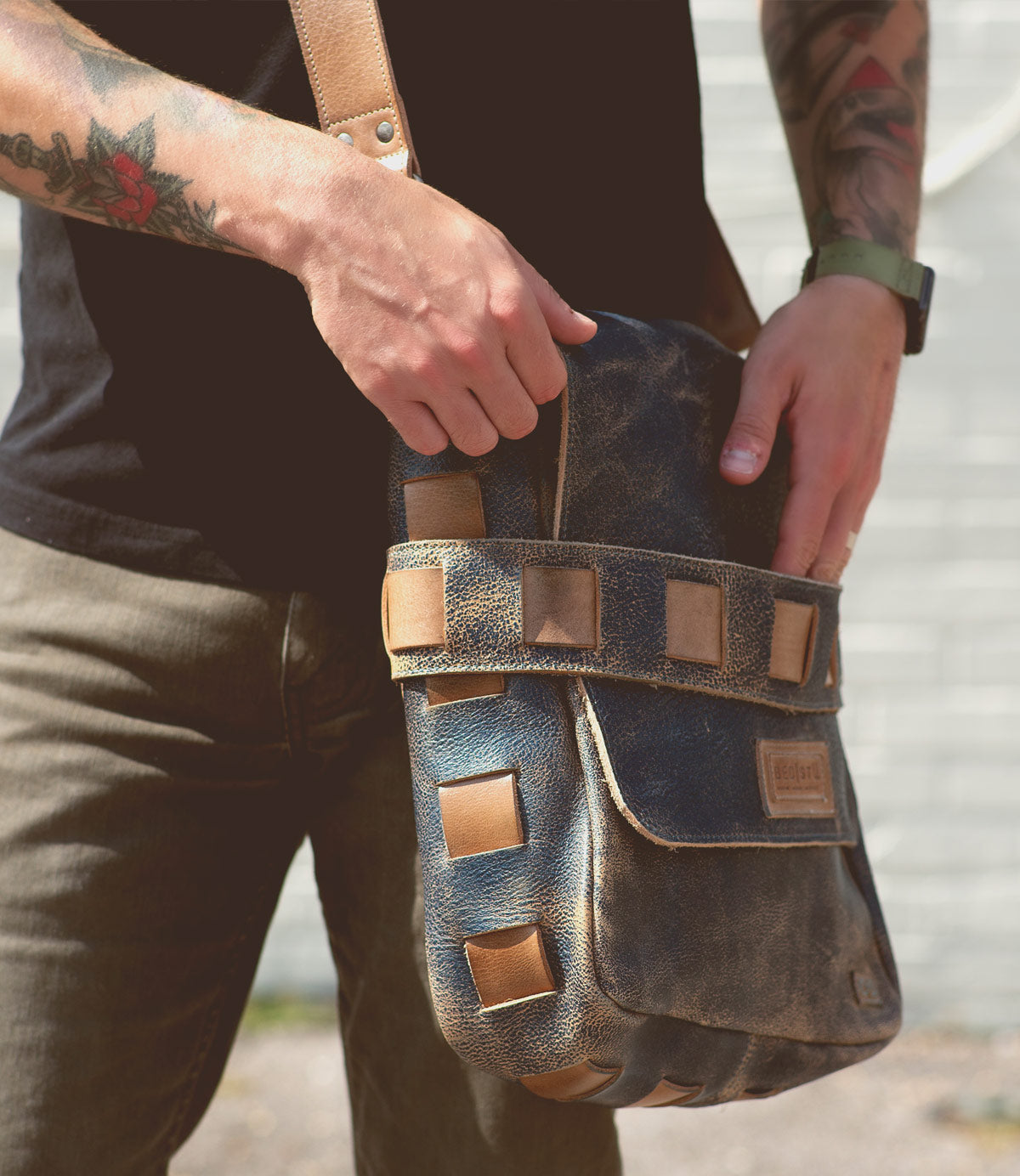 A person with tattoos wearing a black shirt and watch holds the Bed|Stü Kure, a worn brown vegetable tanned leather messenger bag with square accents.