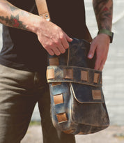A person with tattoos wearing a black shirt and watch holds the Bed|Stü Kure, a worn brown vegetable tanned leather messenger bag with square accents.