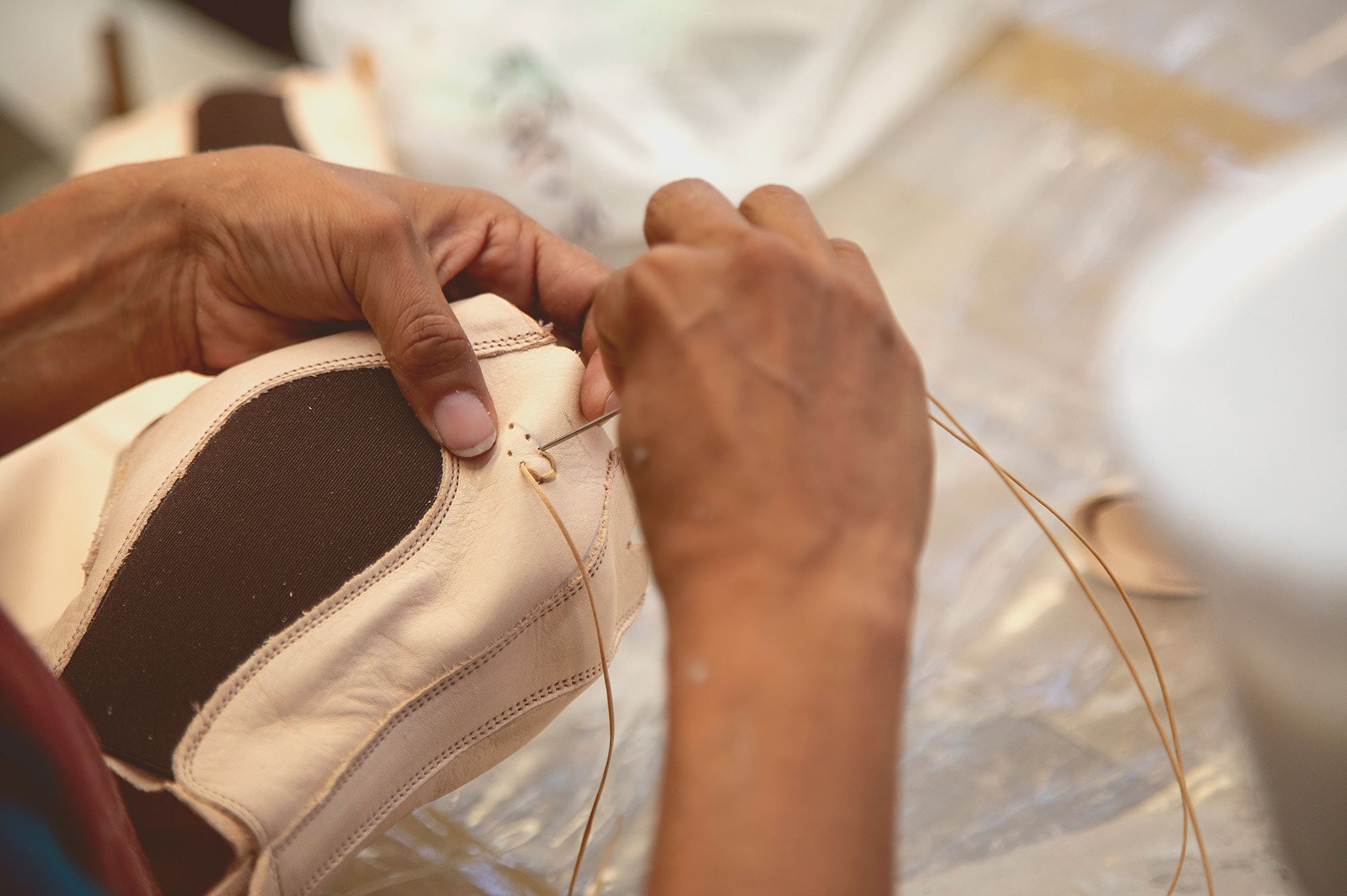 Hands hand-stitching a light leather shoe upper with a needle and tan thread.