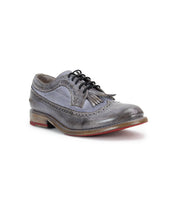 The Lita K by Bed Stu features a gray leather brogue style in a derby shape with a wingtip toe, fringe, black laces, and a red-soled wooden heel, showcased on a white background.