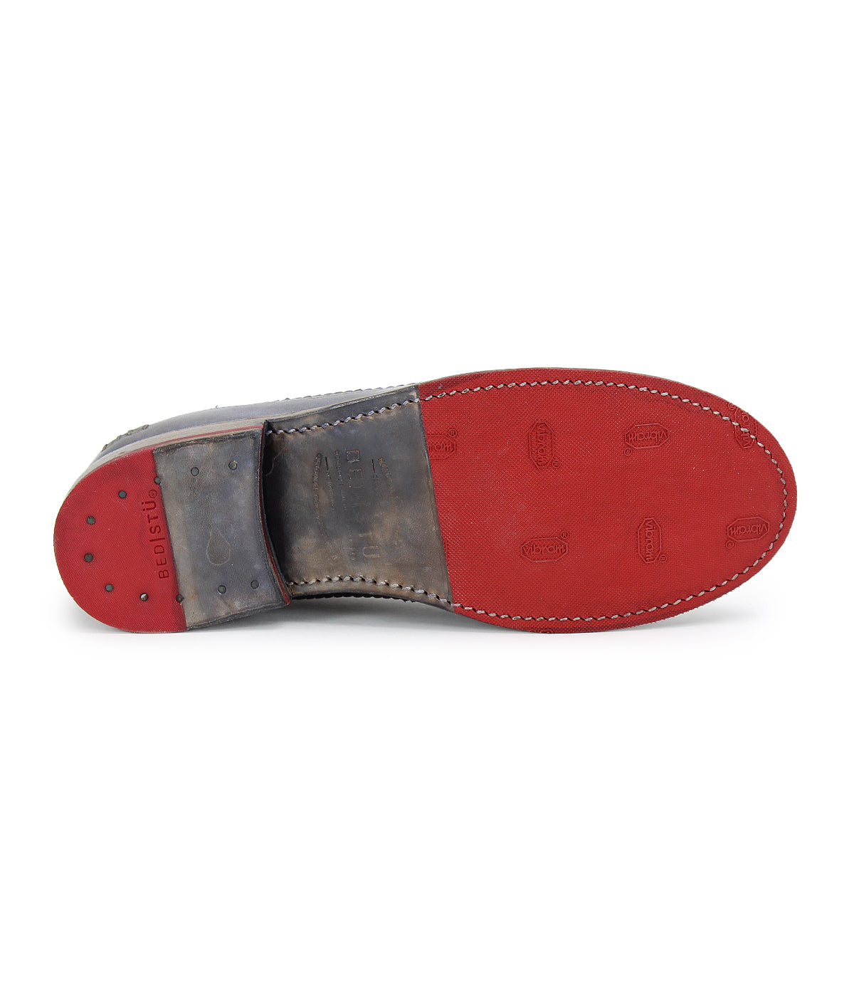 The Bed|Stü Lita K leather dress shoe is shown from the bottom, highlighting its red sole with rubber grips and visible edge stitching, while classic brogue details along the wingtip toe add a touch of sophistication.