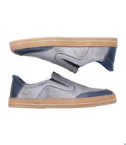 Introducing the Bed|Stü Lanius: a pair of gray slip-on sneakers featuring tan soles, navy blue accents on the heel and toe, and visible stitching. These shoes showcase a timeless silhouette enhanced with modern color blocking, beautifully displayed side by side on a white background.