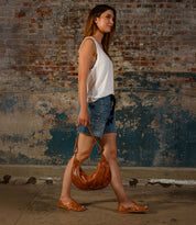 A woman in a sleeveless white top and denim shorts walks past a weathered brick wall, holding a brown leather bag and wearing the Lauren II huarache sandals by Bed|Stü—a beautiful example of handcrafted footwear.
