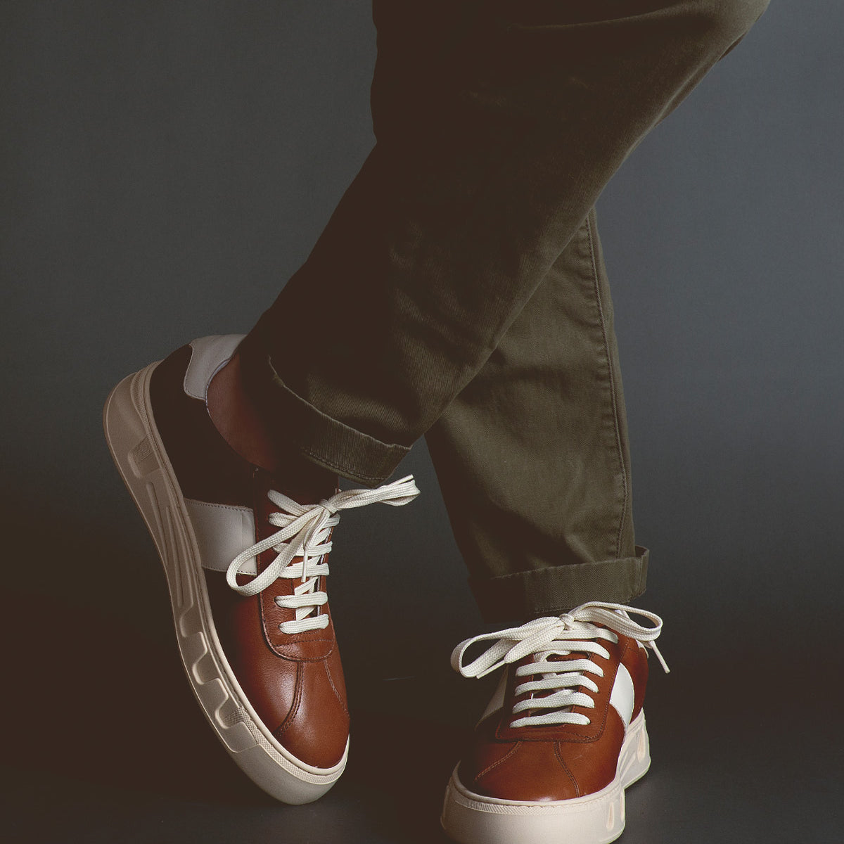 A person wearing olive green pants and Bed|Stü’s Law luxury leather sneakers in brown, white, and black stands with legs crossed against a dark background, highlighting the timeless style and refined Italian craftsmanship of these sneakers.