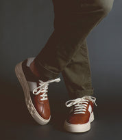A person wearing olive green pants and Bed|Stü’s Law luxury leather sneakers in brown, white, and black stands with legs crossed against a dark background, highlighting the timeless style and refined Italian craftsmanship of these sneakers.
