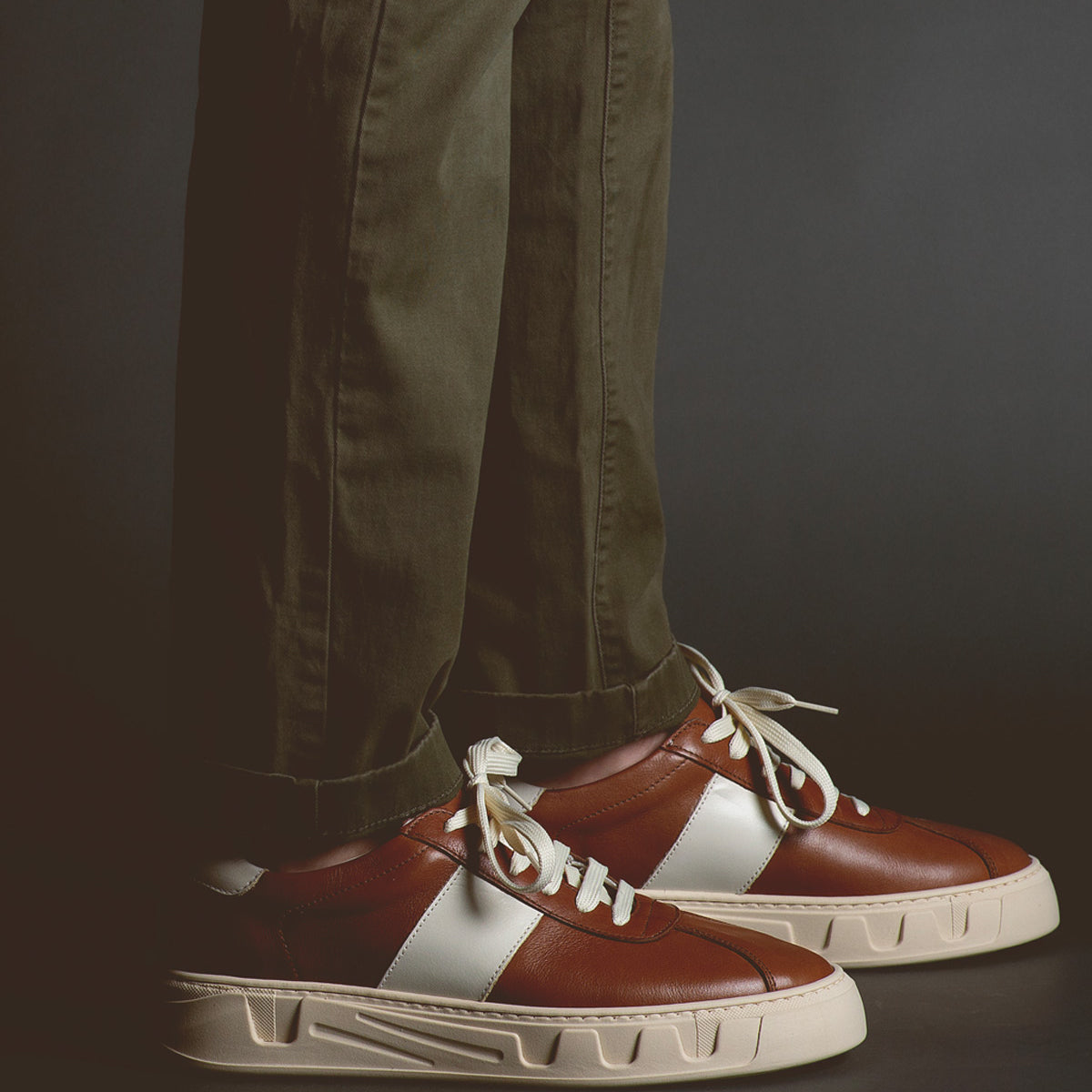 A person wearing olive-green pants and Bed|Stü’s Law luxury leather sneakers—featuring white laces and distinctive white side stripes—stands against a dark backdrop, embodying timeless sneaker style and refined Italian craftsmanship.