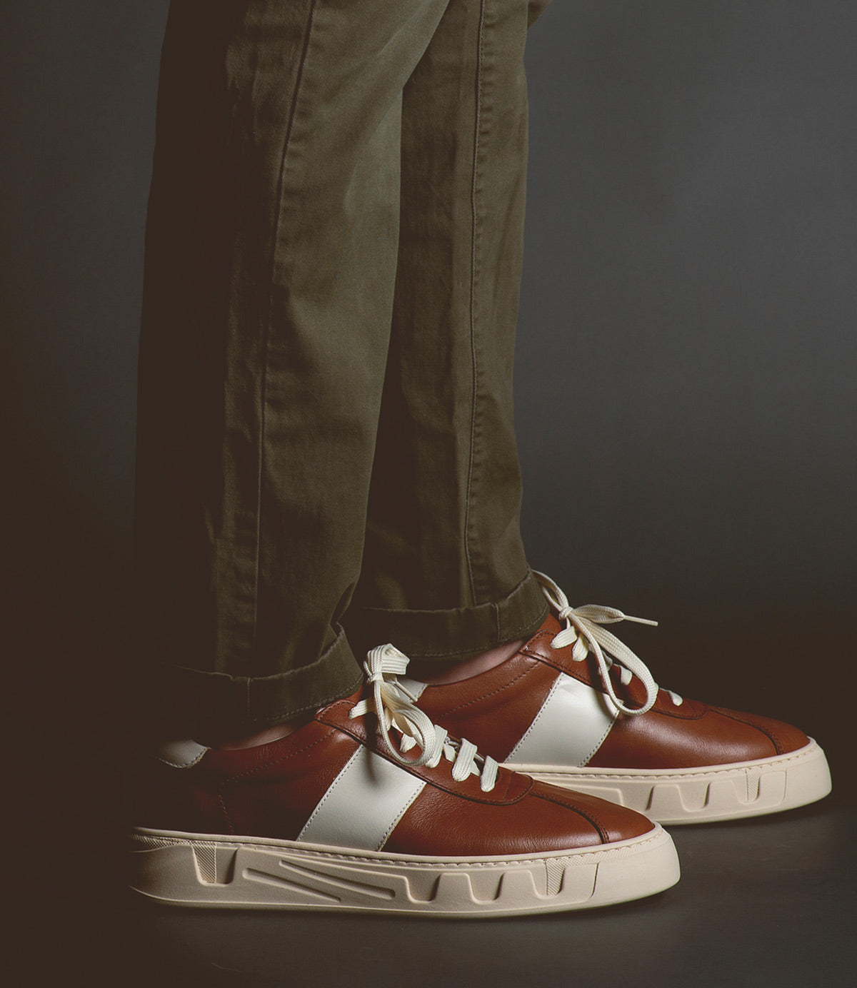 A person wearing olive-green pants and Bed|Stü’s Law luxury leather sneakers—featuring white laces and distinctive white side stripes—stands against a dark backdrop, embodying timeless sneaker style and refined Italian craftsmanship.