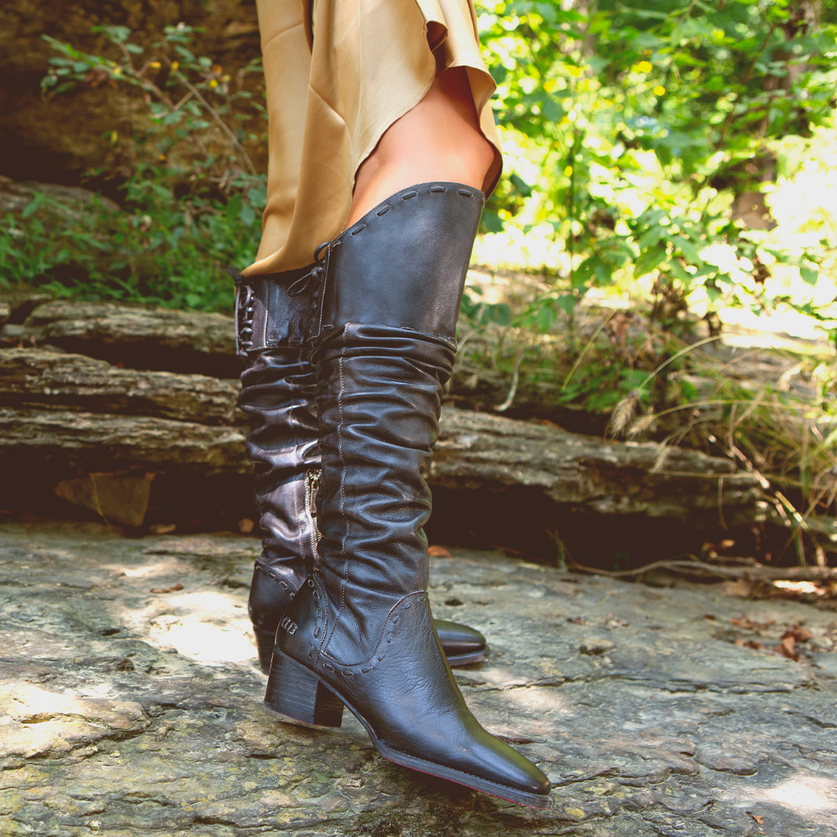 A person stands on rocky ground outdoors amid green foliage, wearing the Leilani beige dress with elegant ruching and Bed|Stü black heeled knee-high stand-out boots.
