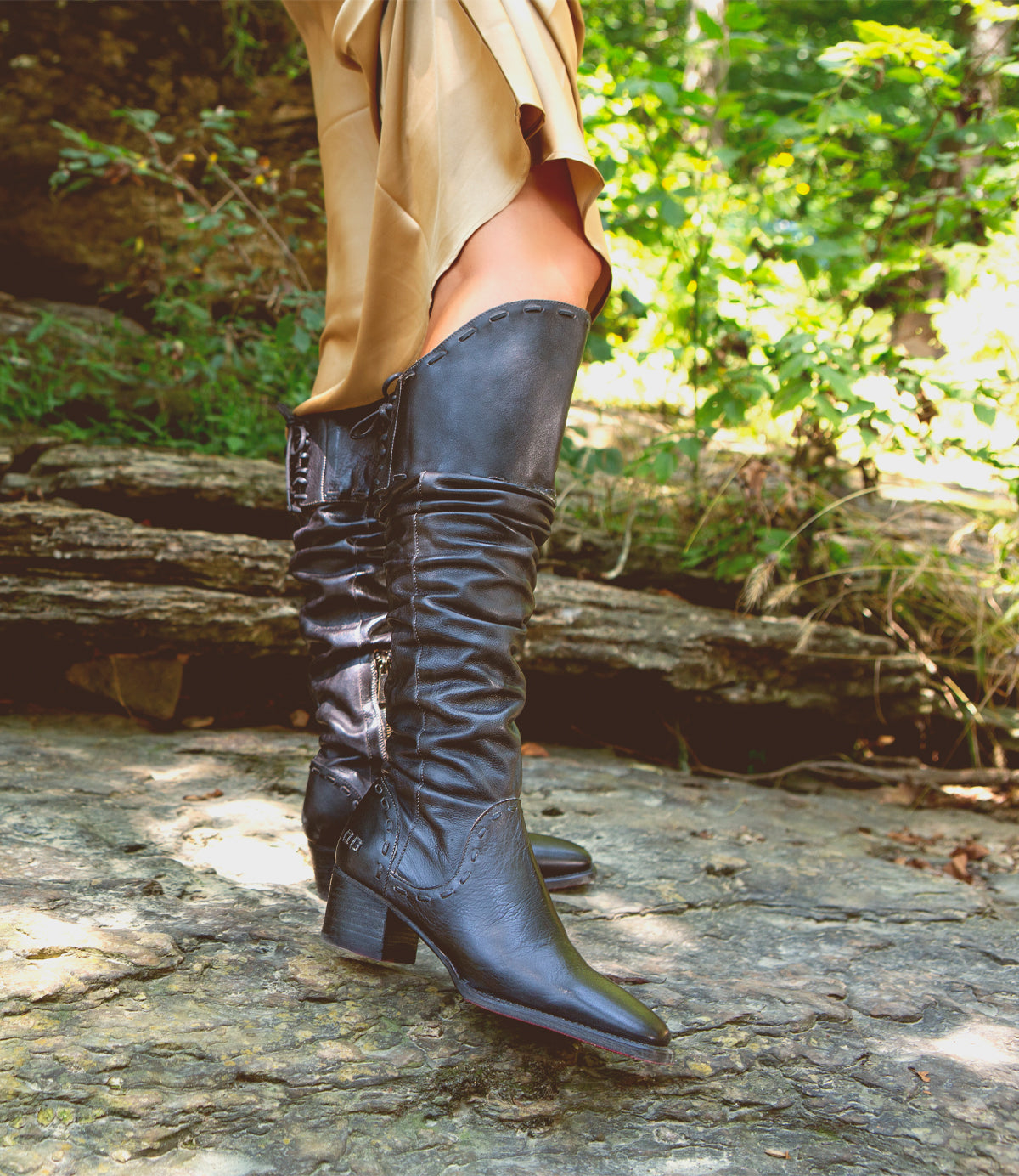 A person stands on rocky ground outdoors amid green foliage, wearing the Leilani beige dress with elegant ruching and Bed|Stü black heeled knee-high stand-out boots.