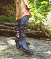 A person stands on rocky ground outdoors amid green foliage, wearing the Leilani beige dress with elegant ruching and Bed|Stü black heeled knee-high stand-out boots.
