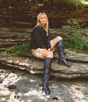 A woman with long blonde hair sits on layered rocks outdoors, wearing a black jacket, a tan skirt with elegant ruching, and tall black Leilani boots by Bed|Stü.
