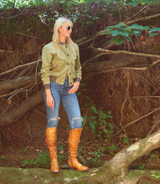 A person stands outdoors in front of exposed tree roots, wearing sunglasses, a green jacket, blue ripped jeans, and standout Bed|Stü Leilani boots featuring elegant ruching.