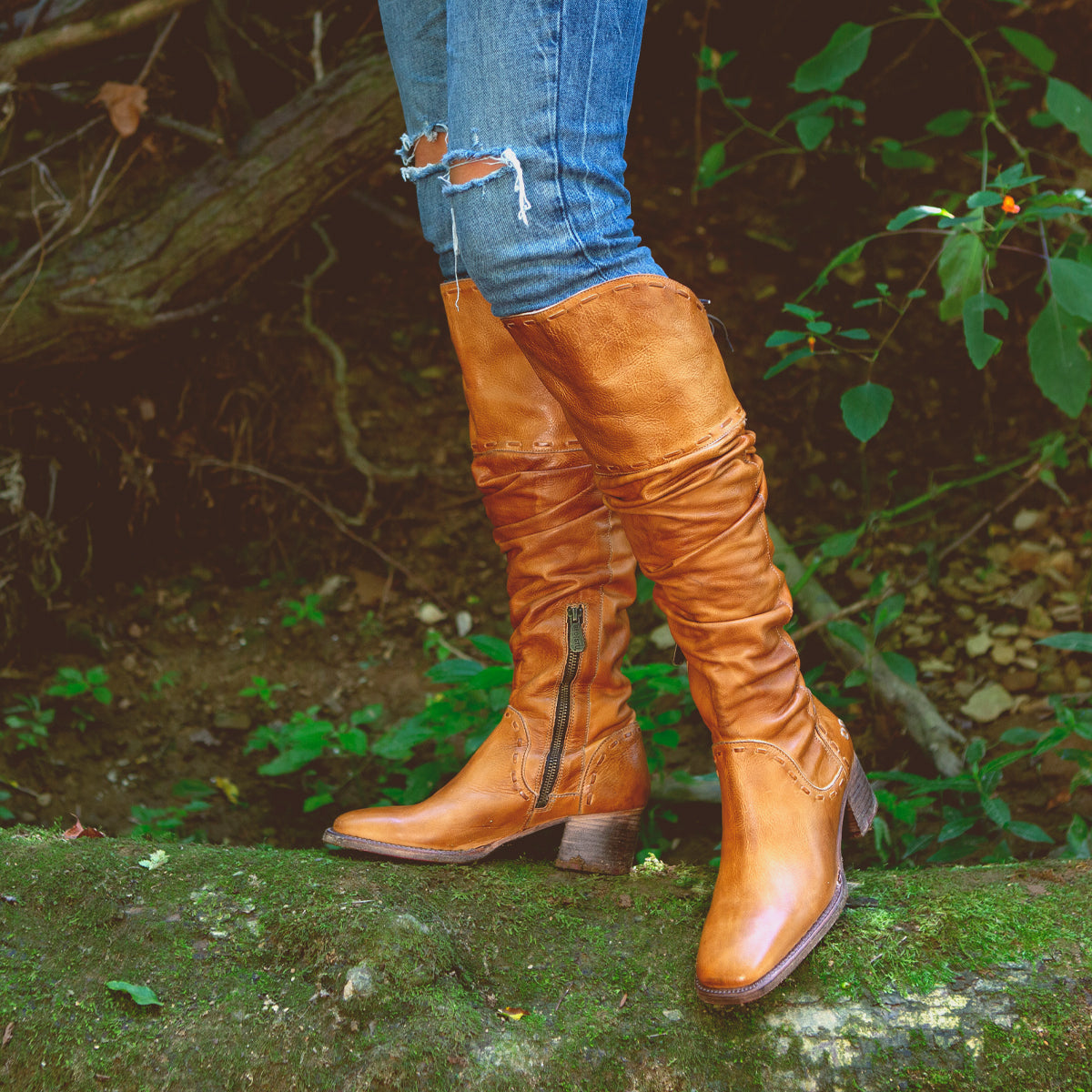 Wearing the Bed|Stü Leilani tan knee-high leather boots, featuring elegant ruching and stylish back laces, someone stands on a mossy log outdoors. With ripped blue jeans tucked in and lush green foliage behind them, this is truly a standout moment for these remarkable boots.
