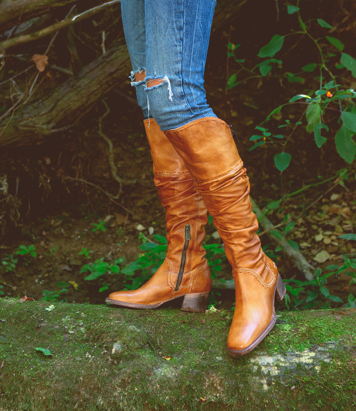 Wearing the Bed|Stü Leilani tan knee-high leather boots, featuring elegant ruching and stylish back laces, someone stands on a mossy log outdoors. With ripped blue jeans tucked in and lush green foliage behind them, this is truly a standout moment for these remarkable boots.
