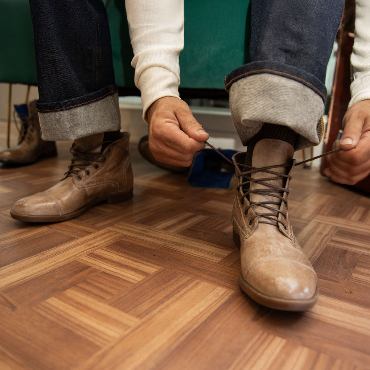 A person in rolled-up jeans and a white shirt is sitting and tying the laces of a brown leather Leonardo boot by Bed|Stü, which features a flexible rubber outsole, on a wooden floor.